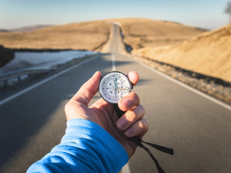 Jemand hält einen Kompass in der Hand. Im Hintergrund eine lange leere Straße, die ins Hinterland führt.