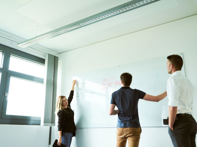 Eine Frau und zwei Männer stehen vor einem Whiteboard und zeichnen einen Verlauf