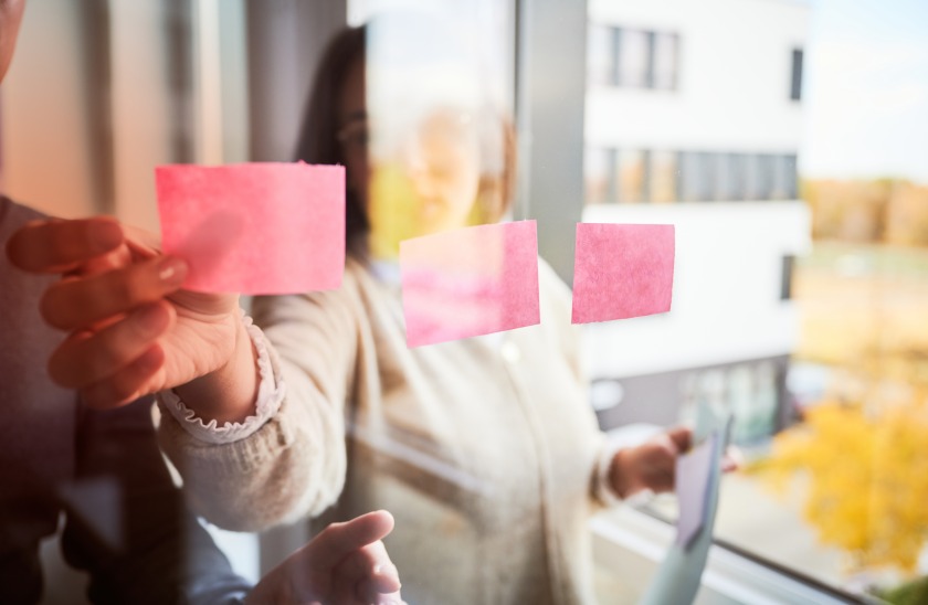 Mitarbeitene kleben Post its an eine Glaswand